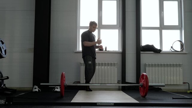 Young male athlete preparing for strength lift on wooden gym platform beside loaded barbell with red bumper plates, standing in bright window backlight during focused training moment