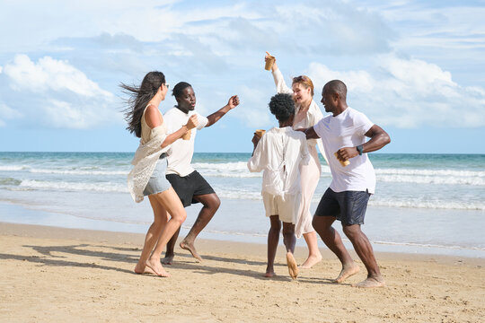 Group of five diverse friends joyfully dancing and celebrating on a sandy beach by the ocean, under a partly cloudy sky