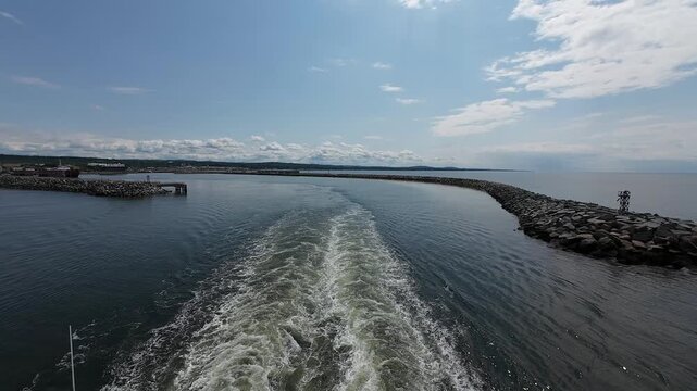 Scenic stern view of a ferry departing Matane Harbor with calm waters, coastal infrastructure, and a bright summer sky.