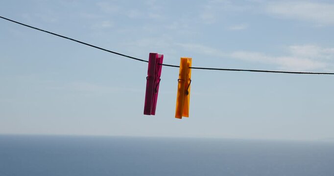 Two colorful clothespins gently sway on a clothesline against a serene blue sky and distant sparkling sea on a beautiful summer day, symbolizing simplicity and freshness.