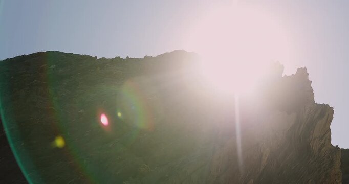 The sun's powerful light emerges behind a rugged mountain silhouette, casting dramatic lens flares across the clear morning sky, symbolizing a new day's dawn and natural beauty.