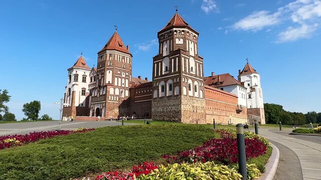 Mir Castle in Belarus. Historic castle with green hill and water reflection, surrounded by trees and clear blue sky, showcasing architectural details in a serene landscape setting