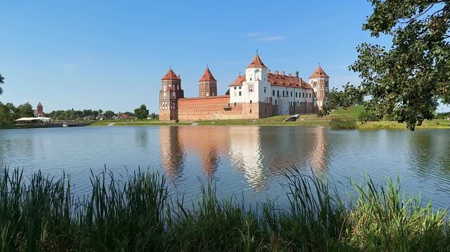 Mir Castle in Belarus. Historic castle with green hill and water reflection, surrounded by trees and clear blue sky, showcasing architectural details in a serene landscape setting