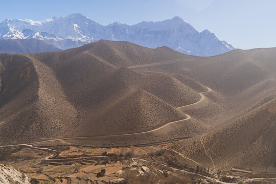 Winding dirt road snakes through the arid mountain slopes of Upper Mustang with snow-capped Himalayan peaks in the background, Nepal.