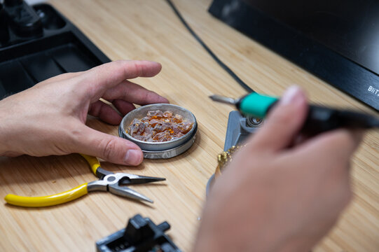 Close-up of a technician's hands holding a metal tin container full of solid pine rosin resin crystals and dipping a hot soldering iron tip into it on a wooden workbench.