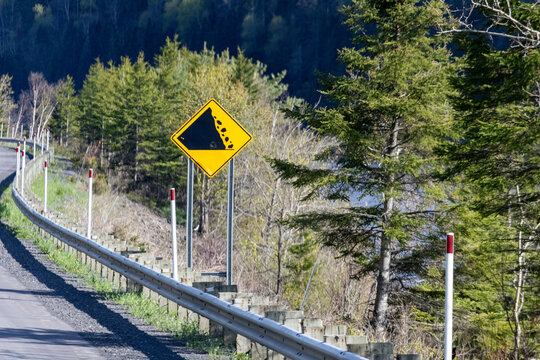 Gaspesie, Canada, 30 May 2026 : A yellow diamond-shaped road sign warns drivers of falling rocks along a winding mountain road with a guardrail.