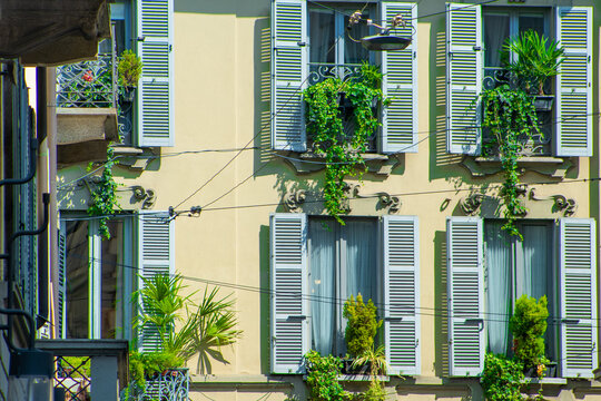 Narrow European Street with Historic Houses and Italian shutters on windows Balcony in Milan. Italy architecture. Green flowers plant in pot.