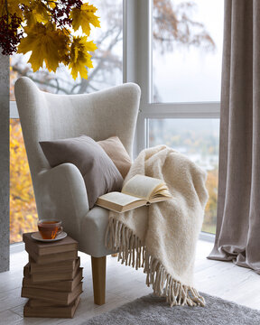 Cozy armchair by large window with autumn leaves above. Stacked books beside warm mug on rustic wood. Soft blanket draped over chair with open book resting