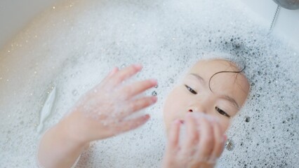 Fototapeta premium A serene young Asian child unwinds in a sudsy bubble bath, a quiet moment of sensory play and wellbeing as part of a daily home hygiene routine.
