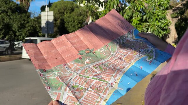 Person examines a folded map outdoors, revealing a colorful city layout with streets and landmarks, surrounded by trees and vehicles in the background
