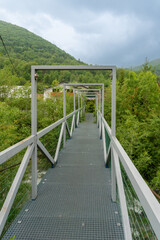 Fototapeta premium A narrow metal footbridge with geometric rectangular frames extends through dense willow vegetation toward buildings beneath forested mountain slopes. Low clouds and mist create an atmospheric landsca