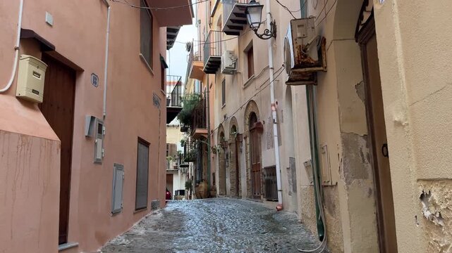 Cefalu, Italy. Narrow cobblestone street lined with pastel-colored buildings, balconies, and outdoor air conditioning units in a quaint Italian town during daylight hours