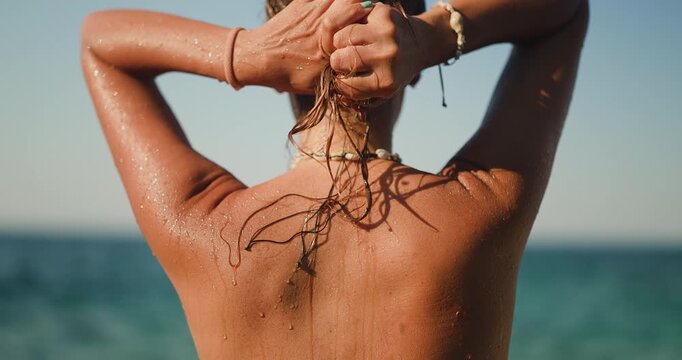 Young woman with wet blonde hair, her tanned back glistening with water drops, enjoying the serene summer sun and ocean breeze after a refreshing swim at the beautiful beach.