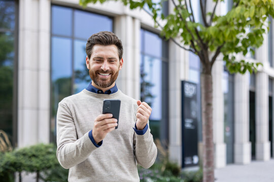 Smiling young man standing on the street near an office building, received a message with better results, rejoices in success