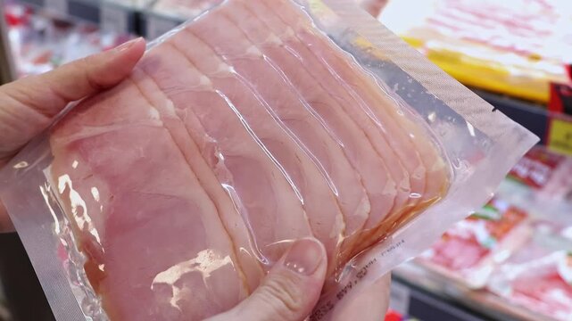 Close up of female hands holding and inspecting plastic vacuum package of sliced ham or pork loin in a supermarket. Customer choosing meat cold cuts in a grocery store. Shopping concept.