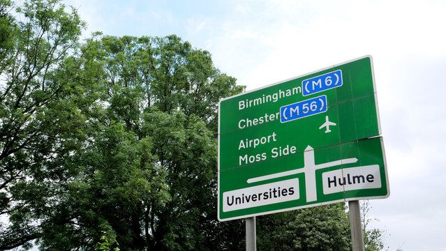 British road sign directing to Birmingham, Chester, Airport, Moss Side, Universities and Hulme against green trees and cloudy sky in Manchester, United Kingdom