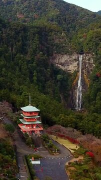 Vertical aerial drone footage of the iconic Seiganto-ji Temple and three-story Nachi Pagoda overlooking the spectacular Nachi Falls in Nachikatsuura, Wakayama Prefecture, Japan. Surrounded by lush mou