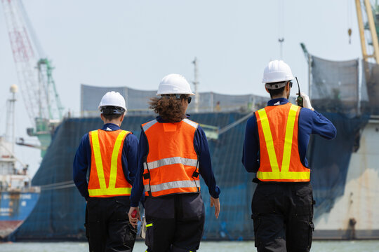 industrial worker walking towards large cargo ship at bustling shipyard wearing white hard hat and bright orange reflective vest feeling determined about upcoming project