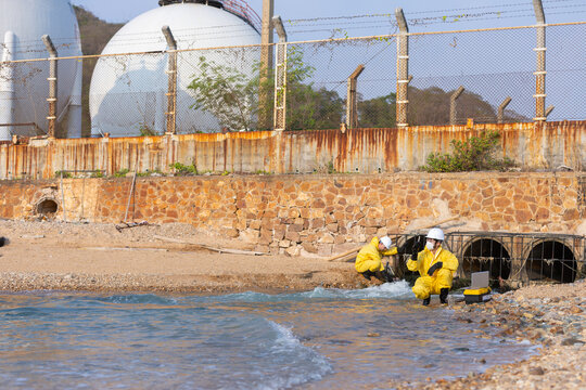 Serious hazmat suit worker team investigates industrial pollution at water beach near large storage tank looking for toxic spill with cautious focused expression during environmental cleanup