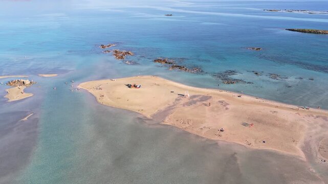 Aerial drone view of clear turquoise sea over a broad rocky reef with a small islet offshore, Elafonissi, Greece