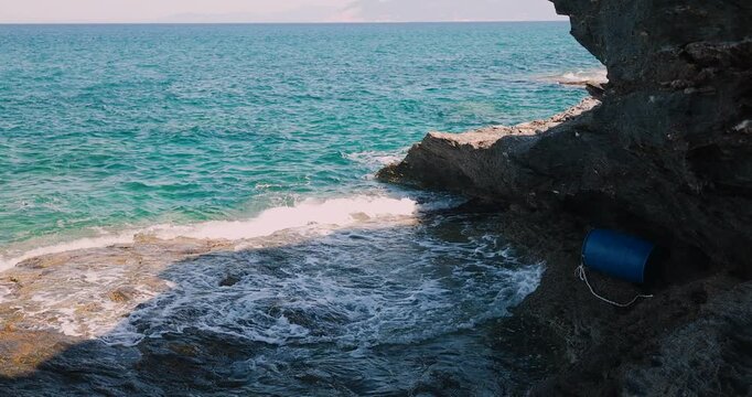 Serene turquoise ocean waves gently crash against rugged coastal rocks on a bright sunny day, with a small blue plastic barrel resting near the water's edge.