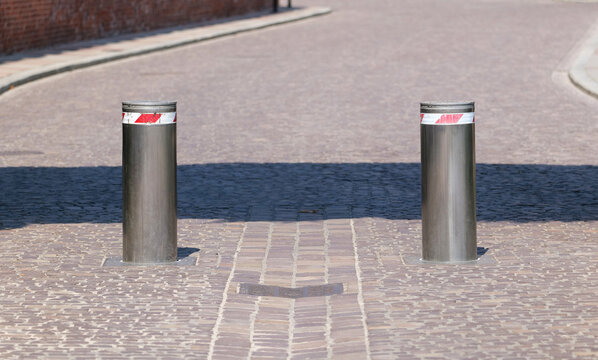 Two raised stainless steel security bollards blocking a cobblestone street, front view of retractable traffic barriers controlling vehicle access in a sunny empty urban road