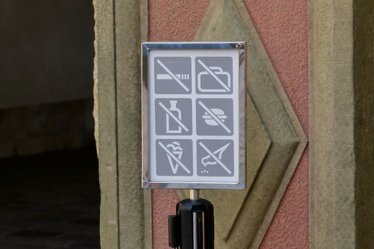 Framed museum prohibition sign showing no smoking, luggage, drinks, food, ice cream or umbrellas, no feeding birds, at a historic entrance, gray pictograms, visitor rules and no people