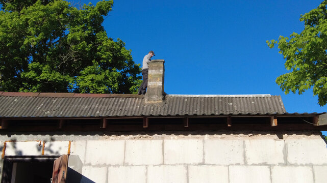 A worker repairs a chimney on the roof of a house under a clear blue sky in a rural area during the day.