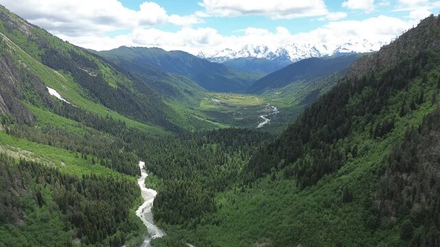 Aerial drone flight at 2,400m toward Becho Valley beneath Mount Ushba's forested slopes, with Dolra River winding below and snow-capped Mount Laila rising under blue sky. Original 4K footage