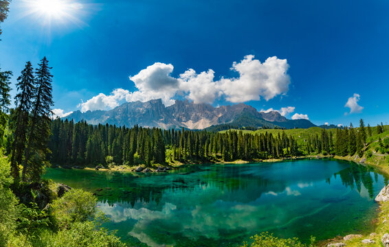 Stunning panoramic view of the emerald green waters of Lake Carezza with Latemar mountain reflection under a bright sunburst, South Tyrol, Dolomites, Italy