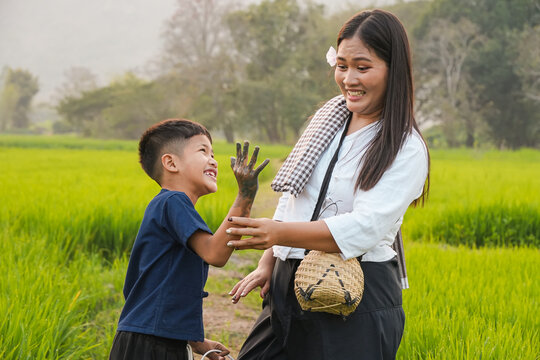 A happy Asian mother and her young son laughing together in a green rice field, playful boy showing his muddy hand during rural family lifestyle outdoor.