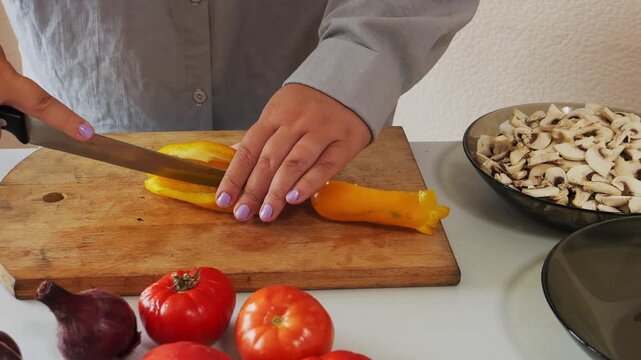 A woman's hands slice yellow peppers on a table with vegetables