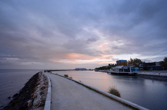A serene waterfront scene at dusk, featuring a calm sea, a rocky jetty,. A boat is docked nearby, with buildings in the background under a cloudy sky.