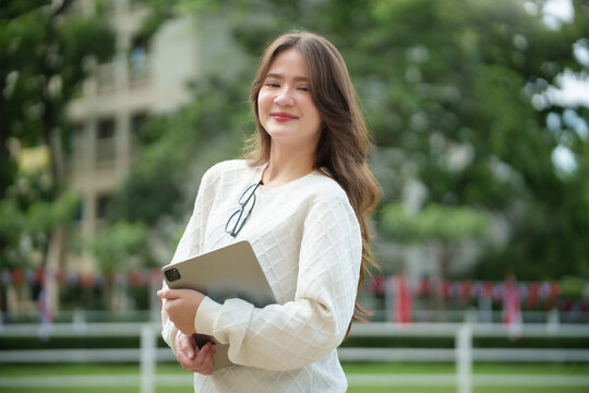 Teenage female university student holding a tablet and smiling at the camera while standing outdoors on campus.