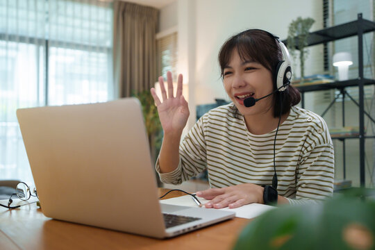 Asian university student waving during an online class at home using a laptop and headset microphone for remote learning.