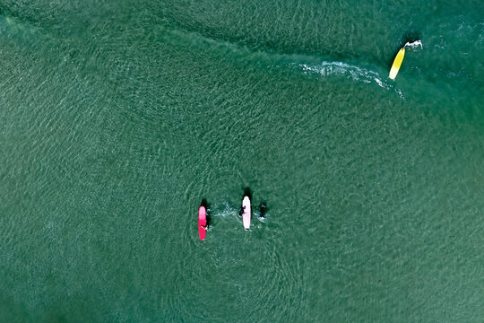 People paddleboard and surf on clear, blue-green ocean water. A sandy beach curves along the shore, with small waves breaking closer to land