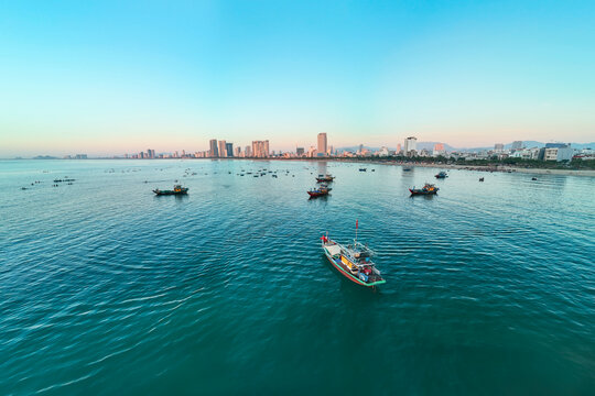 Many colorful fishing boats dot the calm turquoise bay water with a vibrant city skyline and sandy beach visible in Da Nang, Vietnam during early morning