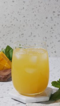 Summer mango drink mocktail with ice and mint on a white coaster, styled in bright sunlight on a terrazzo countertop with fresh mango and clean copy space. A clear rounded glass holds a golden mango