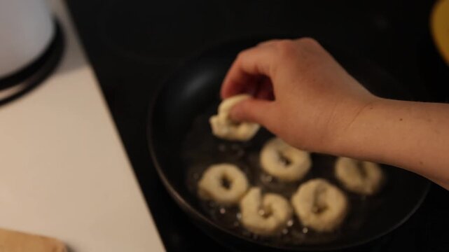 A person is frying donuts in a pan with hot oil, flipping them to achieve a golden‑brown crust, in a kitchen setting, home cooking activity, comfort food preparation.