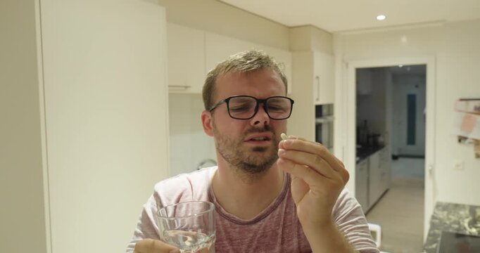 Sweating man taking a tablet with water at home while suffering from migraine, heat stress, and dehydration.