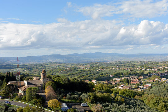 High-angle panoramic view of the rolling hills and green valleys of Umbria, Italy, with a historic stone church and bell tower nestled in autumn foliage under a cloudy sky