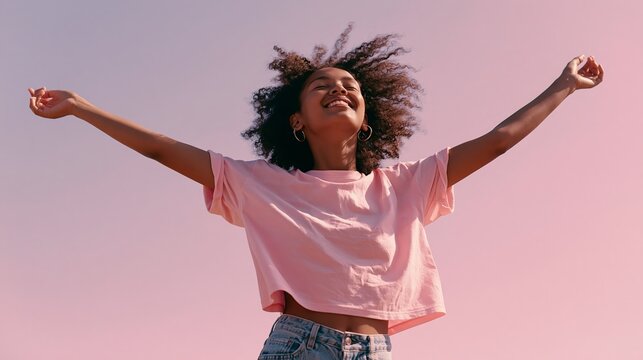 Woman with afro curls joyfully dances in a pink tee and jeans, summer sun, on a pink backdrop.