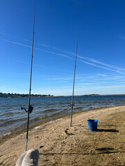 Fototapeta premium Fishing Rods On A Sandy Lake Shore With Bucket And Calm Blue Sky For Relaxing Day