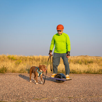 mature senior male is riding one-wheeled electric skateboard with a pitbull dog on a prairie road, late summer scenery
