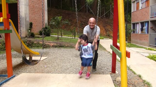 Smiling father pushing his little son on a swing at a residential playground, happy family enjoying outdoor activity together