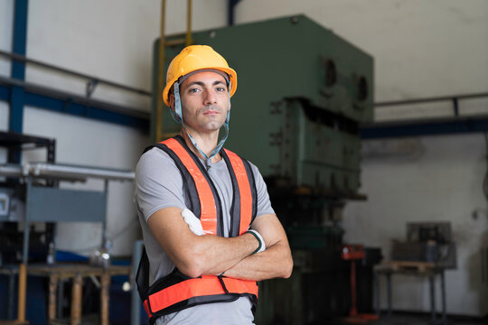 Portrait of male industrial worker standing with crossed arms in factory with blur heavy industry machine background