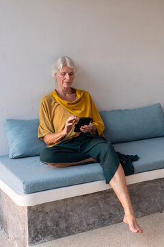 focused senior woman using smartphone, sitting on couch, full body 