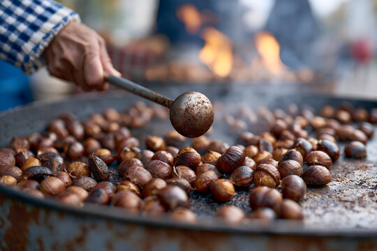 Hand stirs roasted chestnuts over open flame. Chestnuts sizzle, darkening as they cook. Fires glow warmly in background, casting soft light. Texture of nuts and metal pan hints at rustic cooking