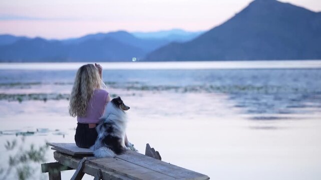 A model and a Shetland Sheepdog sitting on the shore of a lake at dawn.
