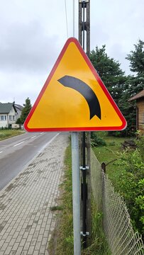Yellow triangular road sign warning of a dangerous curve ahead to the left, European traffic sign on a metal post next to a paved sidewalk and asphalt street in a rural residential area